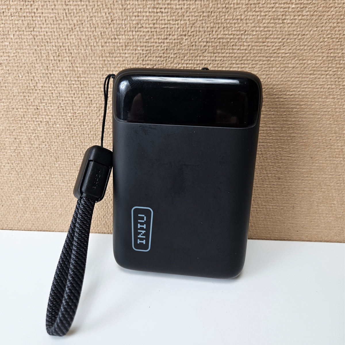 A black INIU portable power bank with a matching wrist strap, standing on a white surface against a textured beige background.