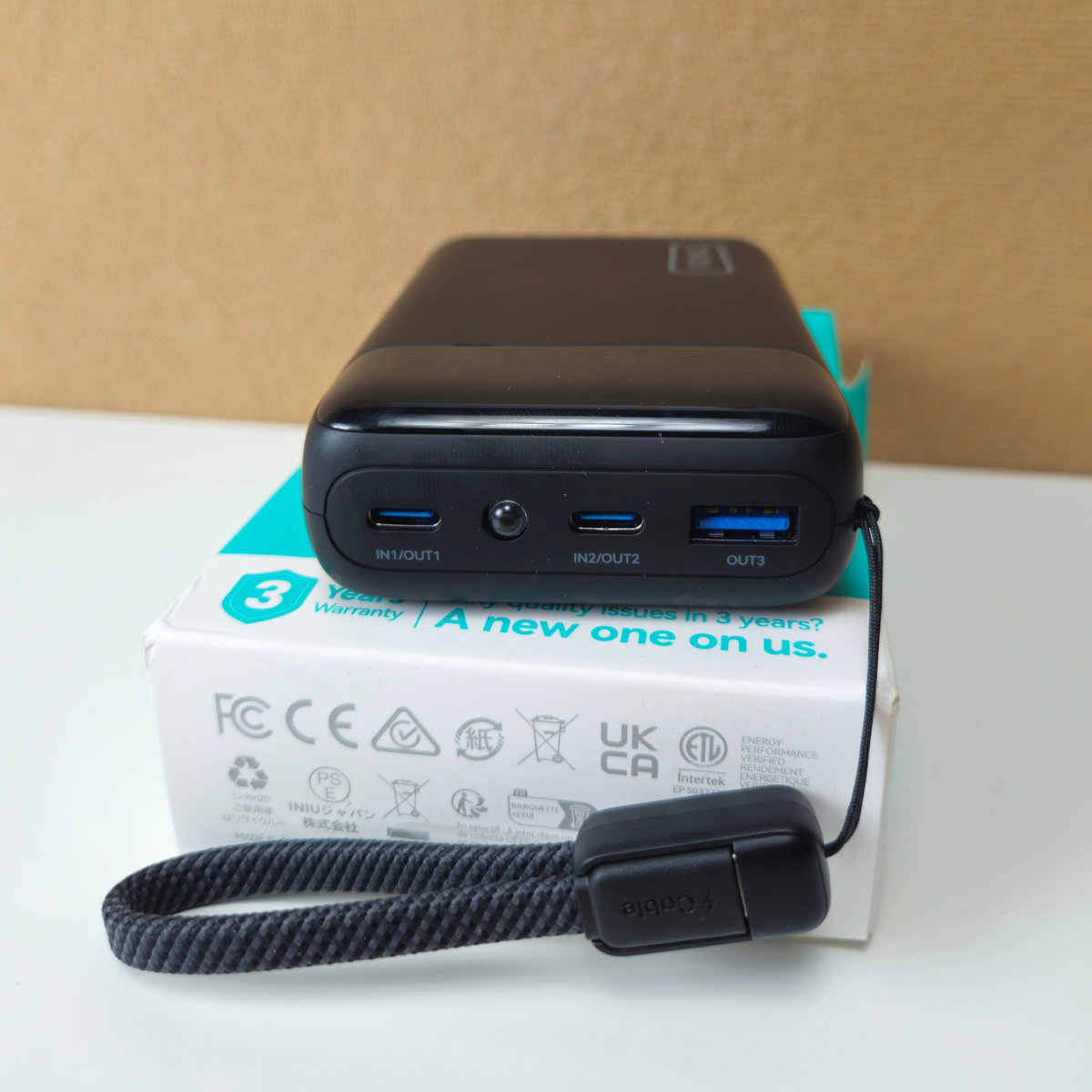 A black portable power bank with three USB-C ports and a headphone jack, resting on top of its white and teal retail box. A black wrist strap with a magnetic clip is attached in front.