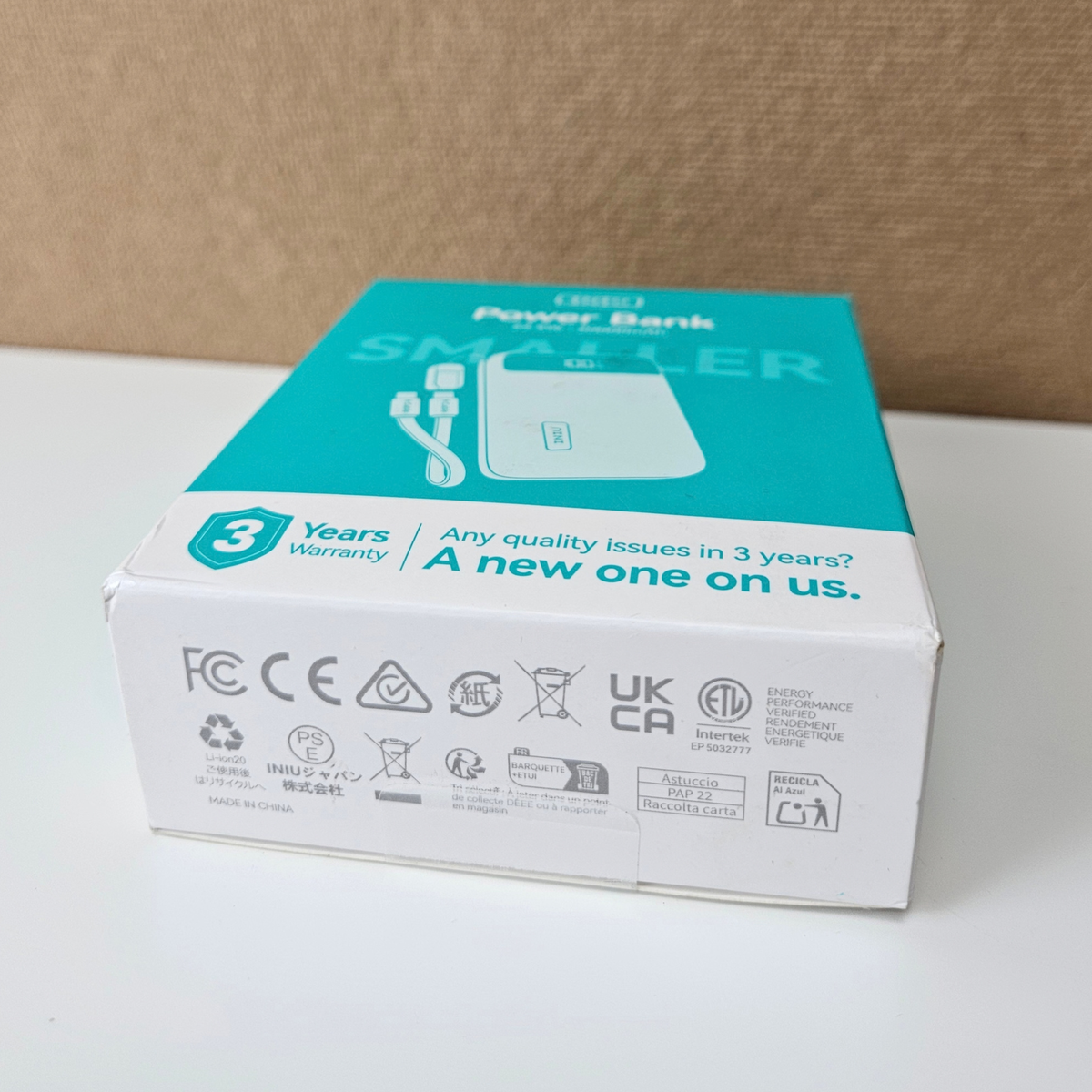 A boxed power bank product package photographed on a white surface against a tan fabric backdrop. The box is mostly white with a bright teal top panel and printed product imagery and regulatory icons on the front and side.