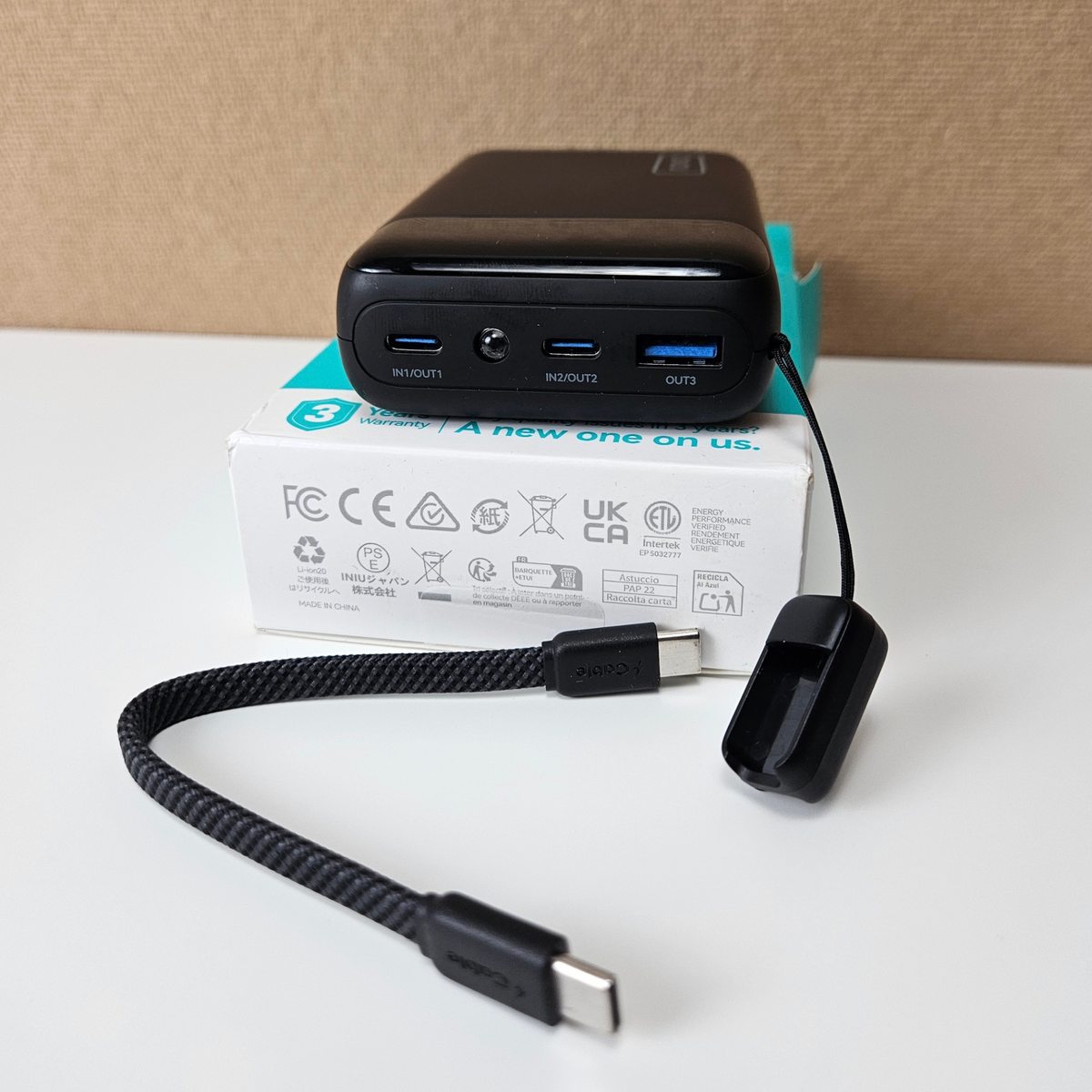 A black portable power bank with USB-C and USB-A ports sits on top of its white product box, with a black braided USB-C cable and a small black accessory placed in front on a white tabletop.