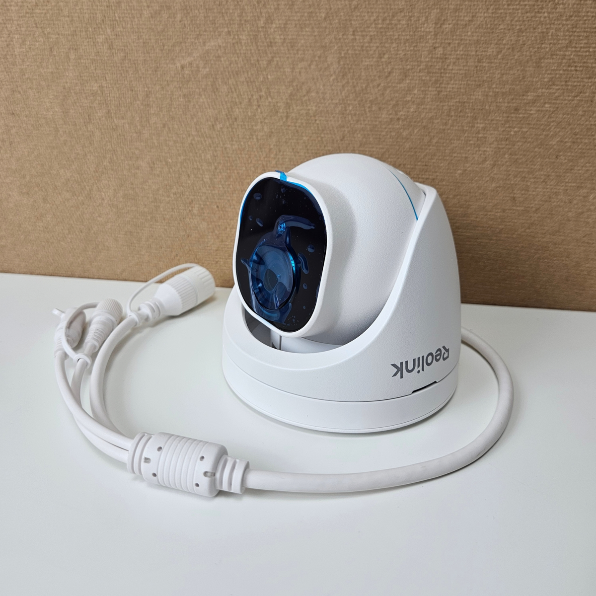 A white Reolink security camera with a coiled white power cable, set against a neutral beige textured background on a white surface.
