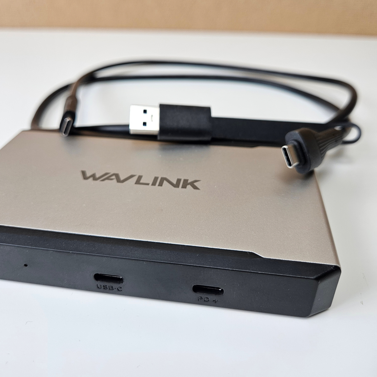 A silver and black Wavlink USB-C hub with a coiled black USB-C cable, photographed on a white surface with a beige background.