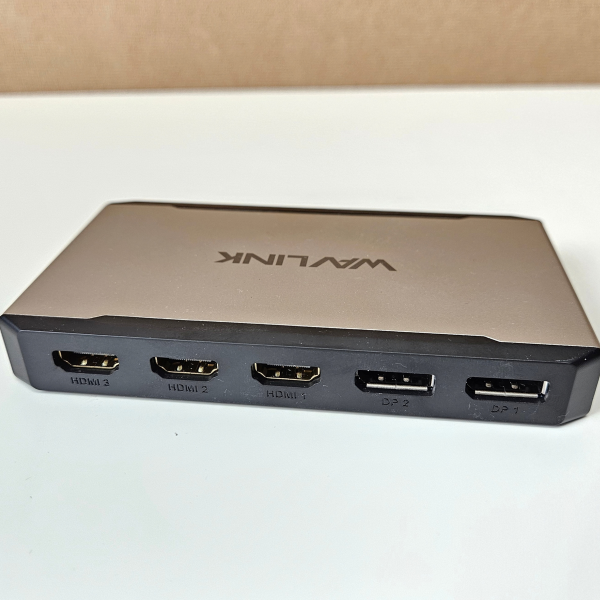 A Wavlink HDMI splitter box with a metallic bronze top and black front ports, shown on a white surface with a brown background.