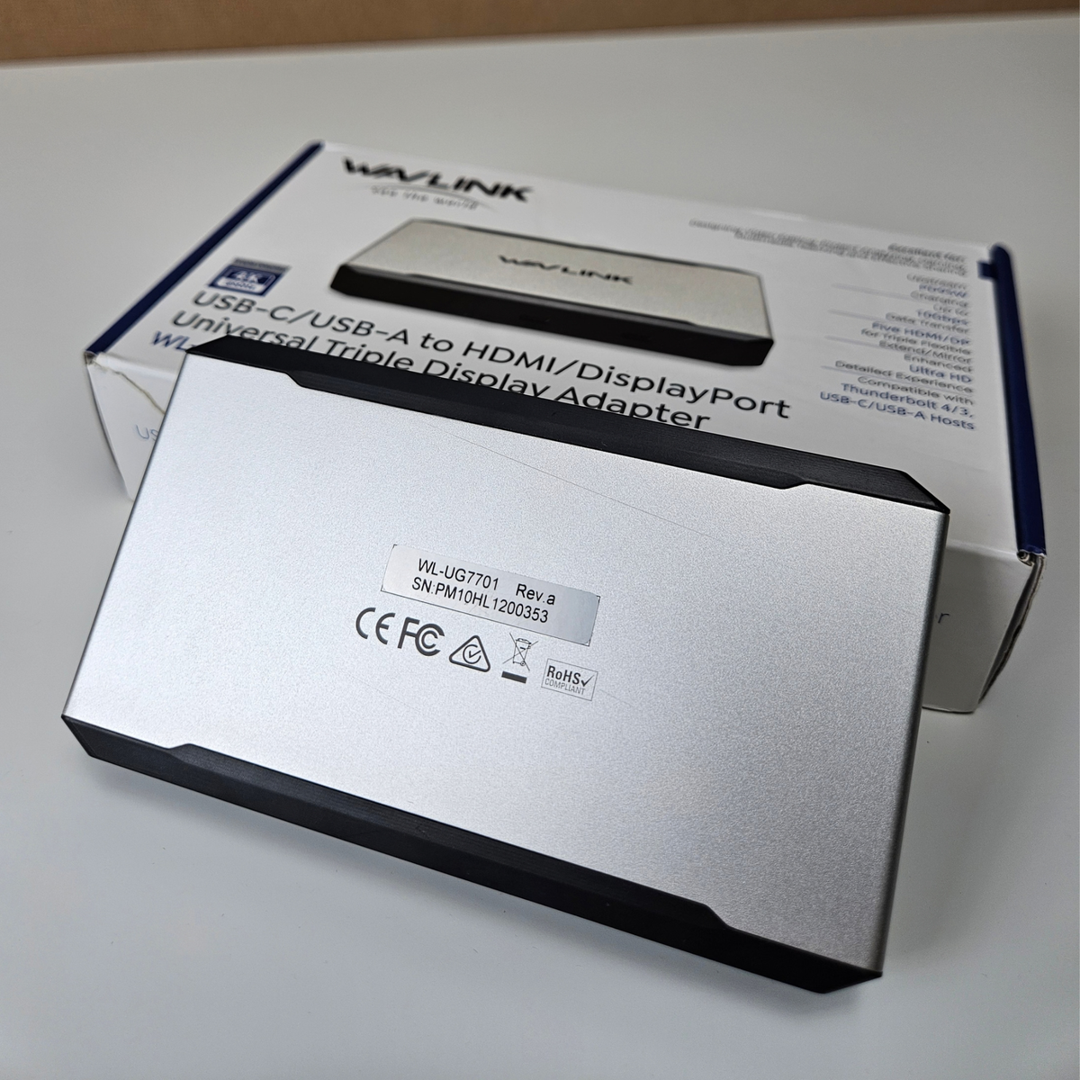 A silver and black Wavlink USB-C to HDMI/displayPort universal triple display adapter in the foreground, with its retail box behind it on a light surface.
