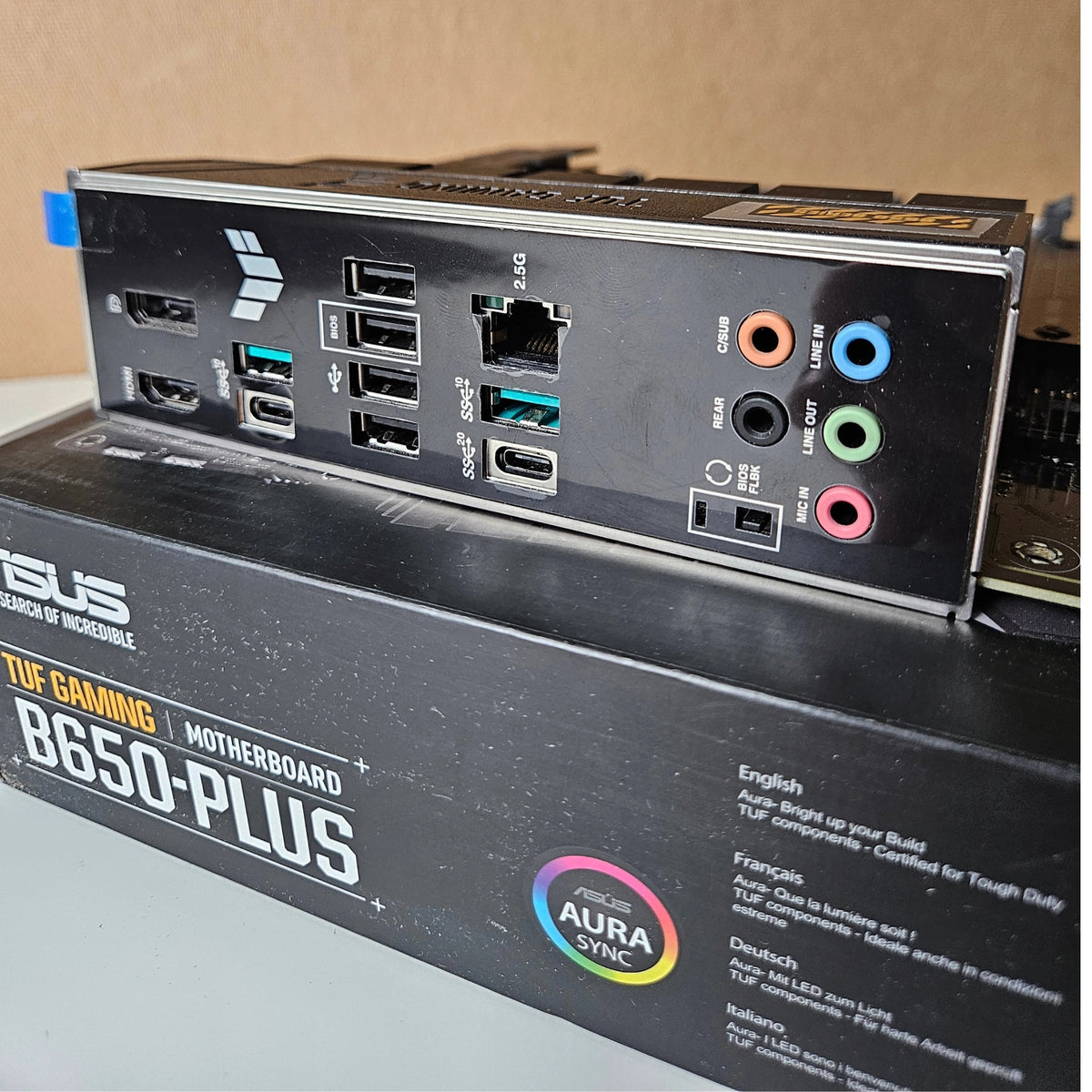 Close-up of the rear I/O panel of an ASUS TUF Gaming B650-PLUS motherboard resting on top of its retail box. The black panel features multiple USB-A and USB-C ports, an Ethernet port, and colorful audio jacks.
