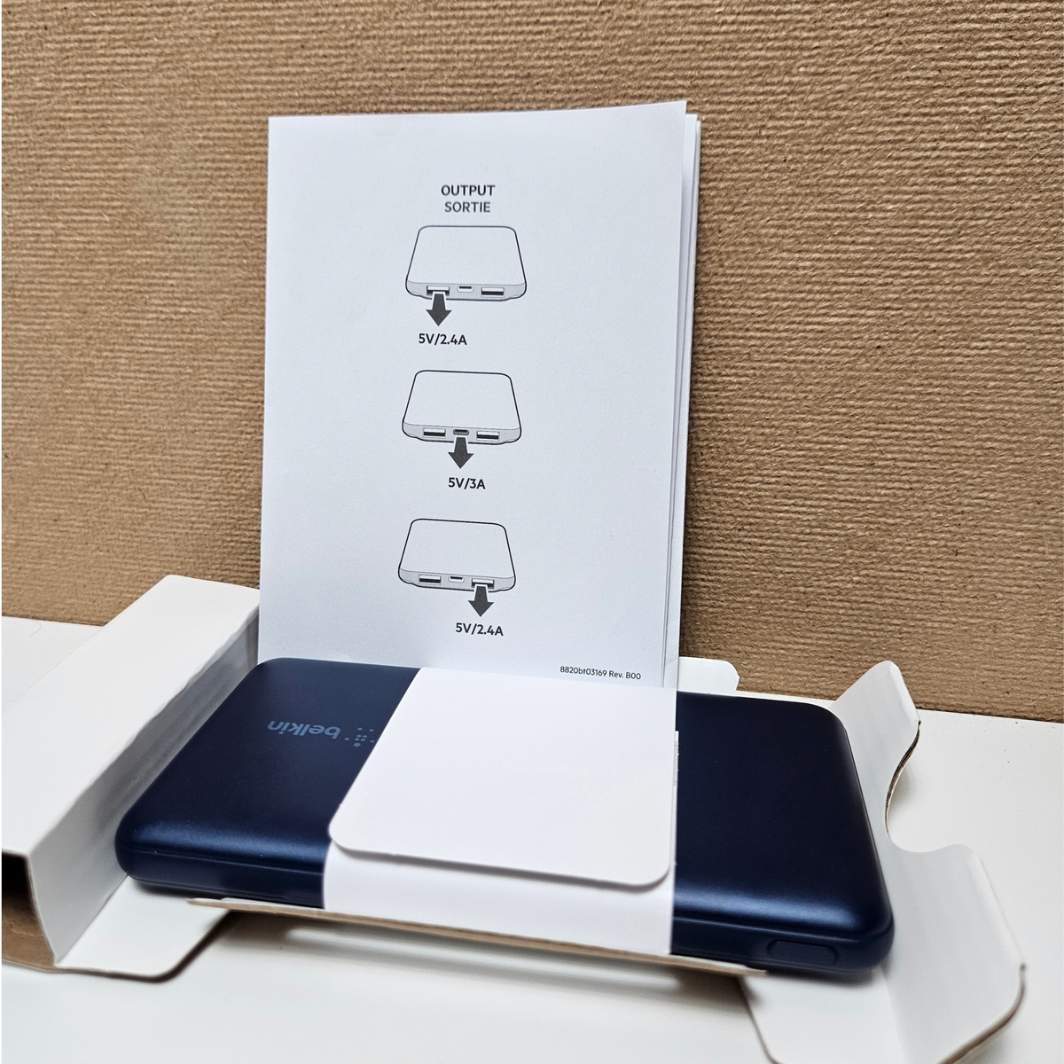 Blue portable power bank in its white box, with a white instruction sheet showing output port configurations standing behind it against a brown textured background.