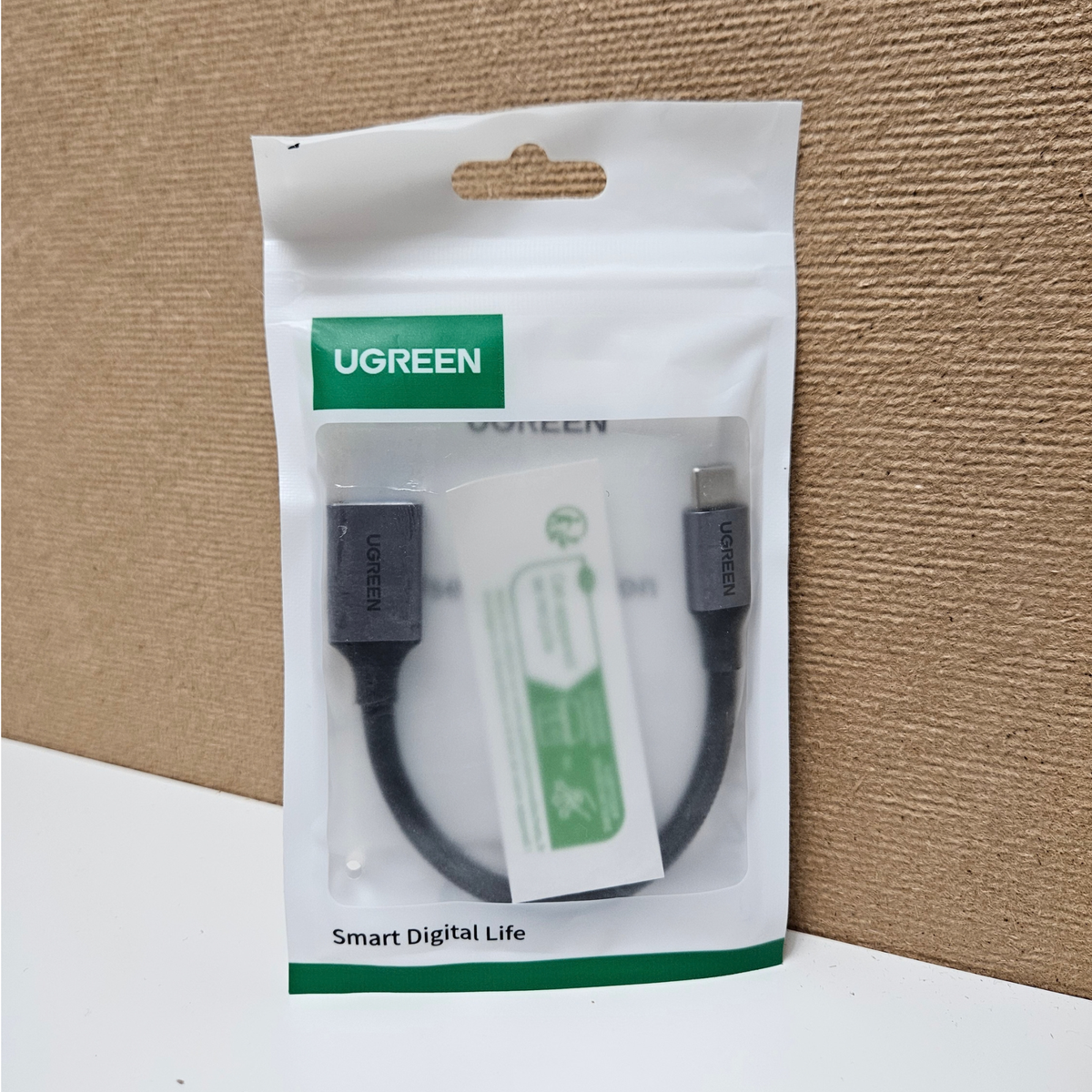 UGREEN cable in its original white and green retail packaging, displayed against a brown corrugated cardboard background.