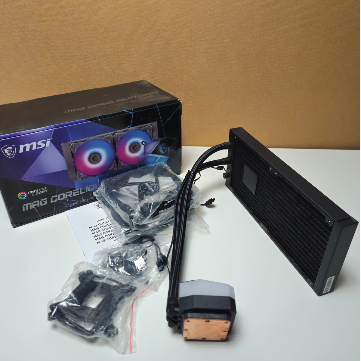 MSI MAG 240mm liquid CPU cooler kit with a black radiator, pump block, and cooling tubes, shown alongside its retail packaging box and mounting hardware on a white surface.