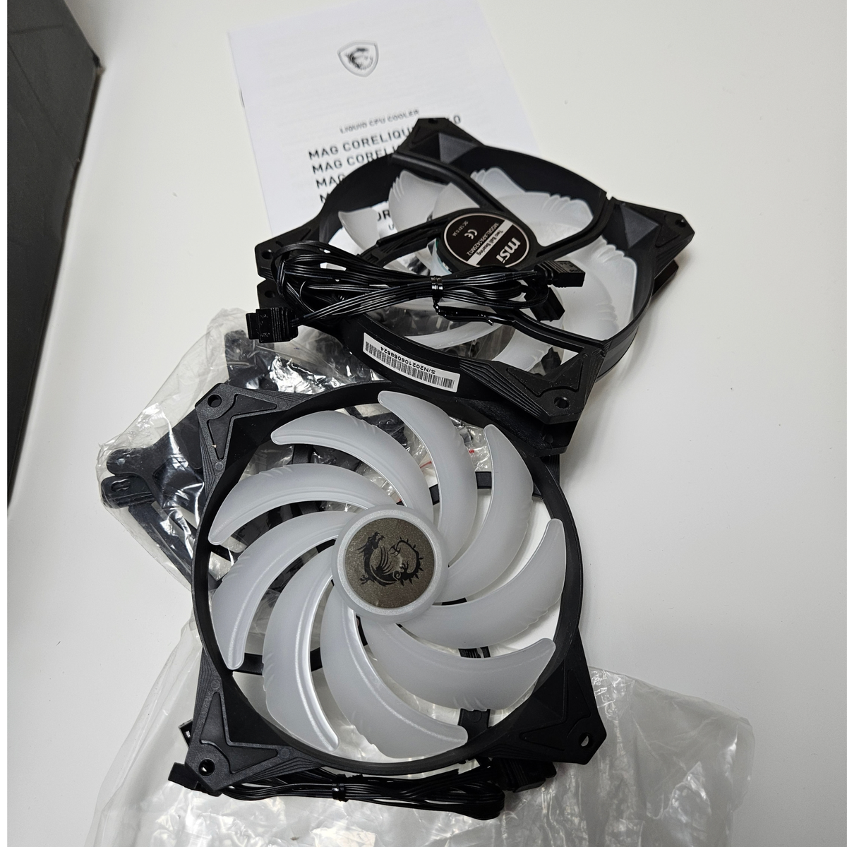 Two MSI  cooling fans, one with white blades and one with black blades, resting on a white surface with their packaging and a manual visible.