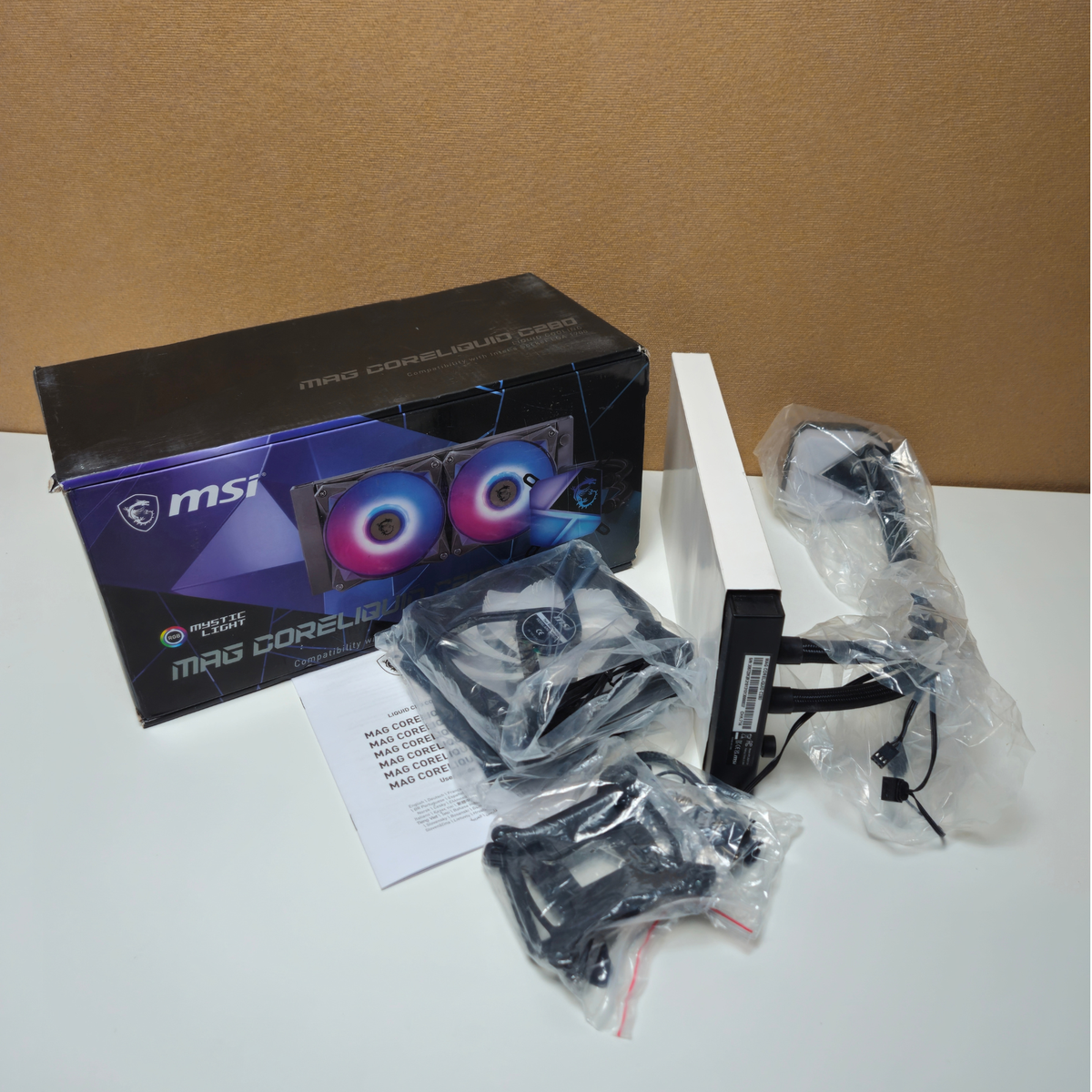 MSI MAG CORELIQUID CPU cooler kit components laid out on a white surface, including the retail box, mounting brackets in plastic bags.