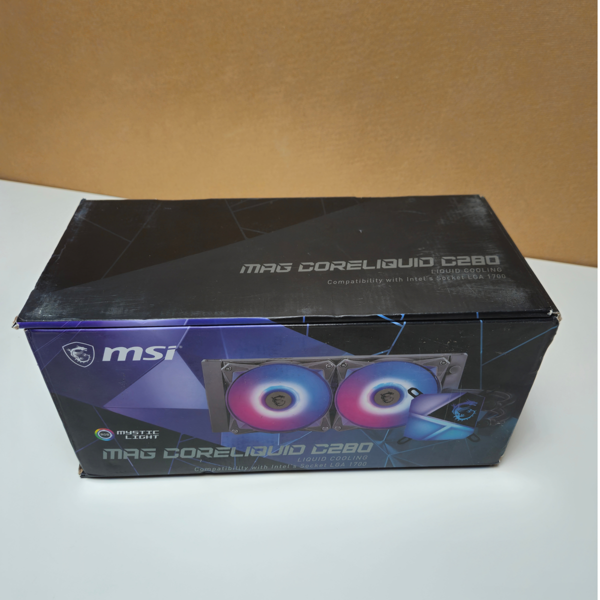MSI MAG Coreliquid C280 liquid CPU cooler retail box, featuring a black and blue design with an illustration of two RGB fans, photographed on a white surface against a brown background.