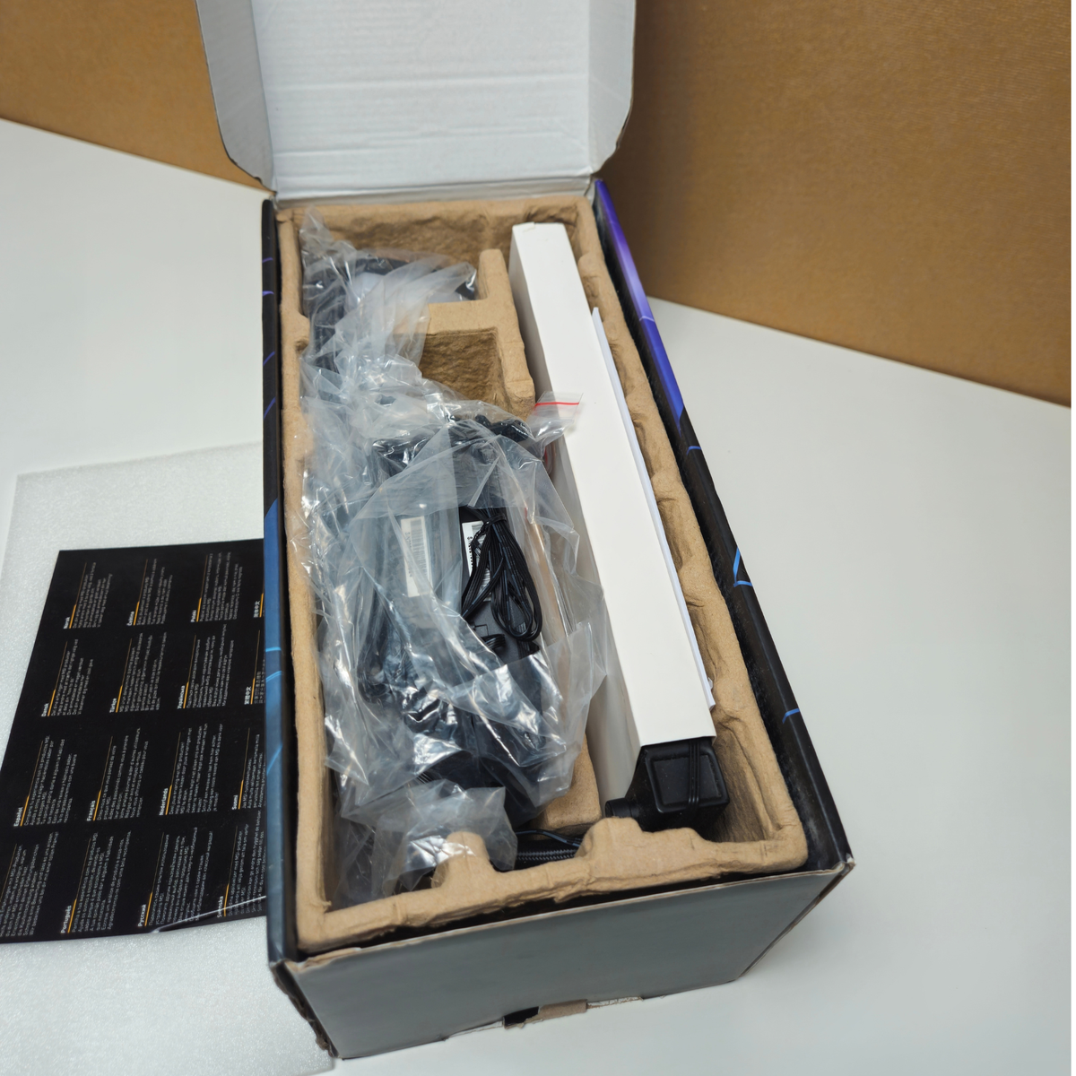 Open product box showing included accessories and packaging inserts on a white tabletop, with a brown cardboard backdrop behind.