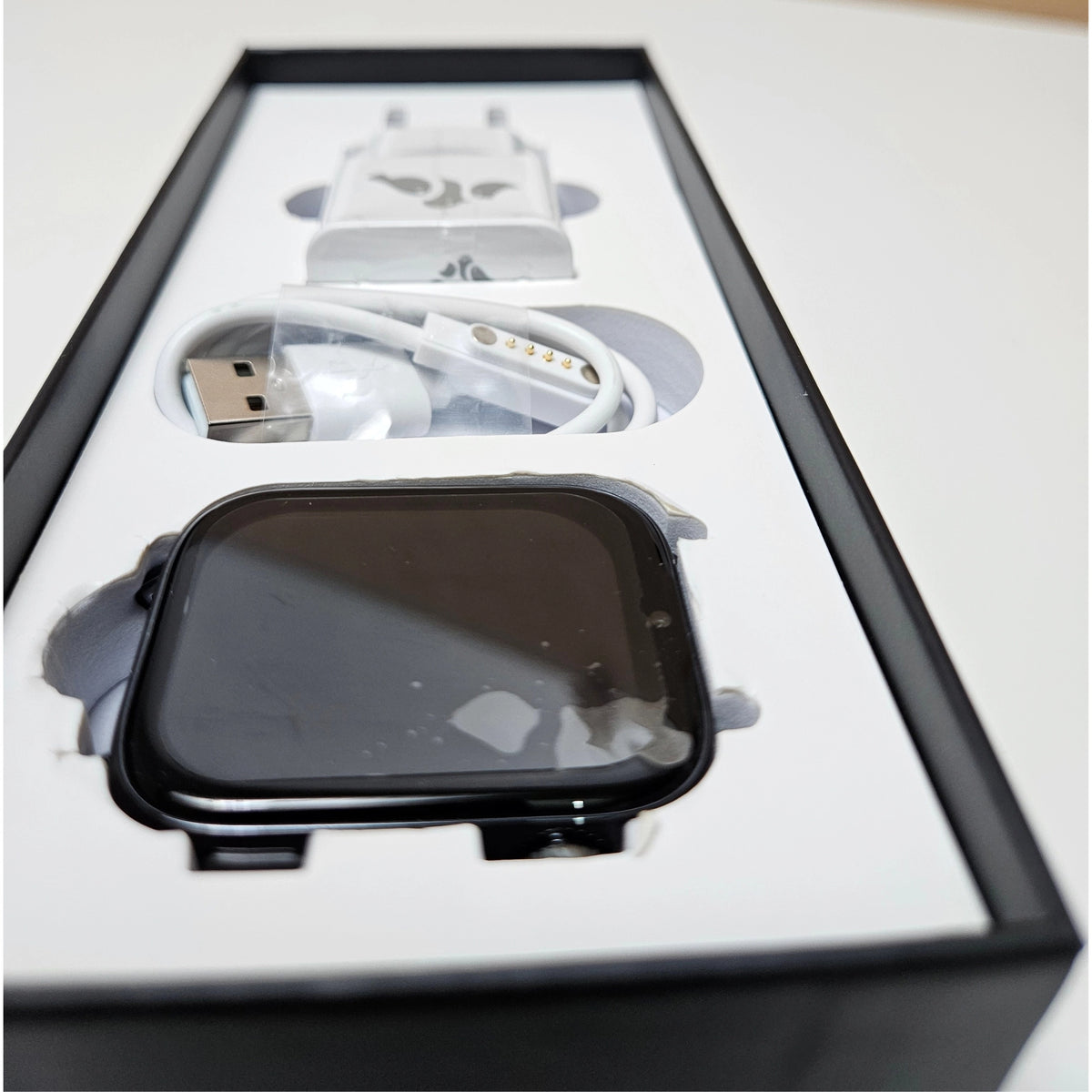 A black smartwatch with a square screen displayed inside its white and black retail box, alongside a white charging cable and a white protective case.