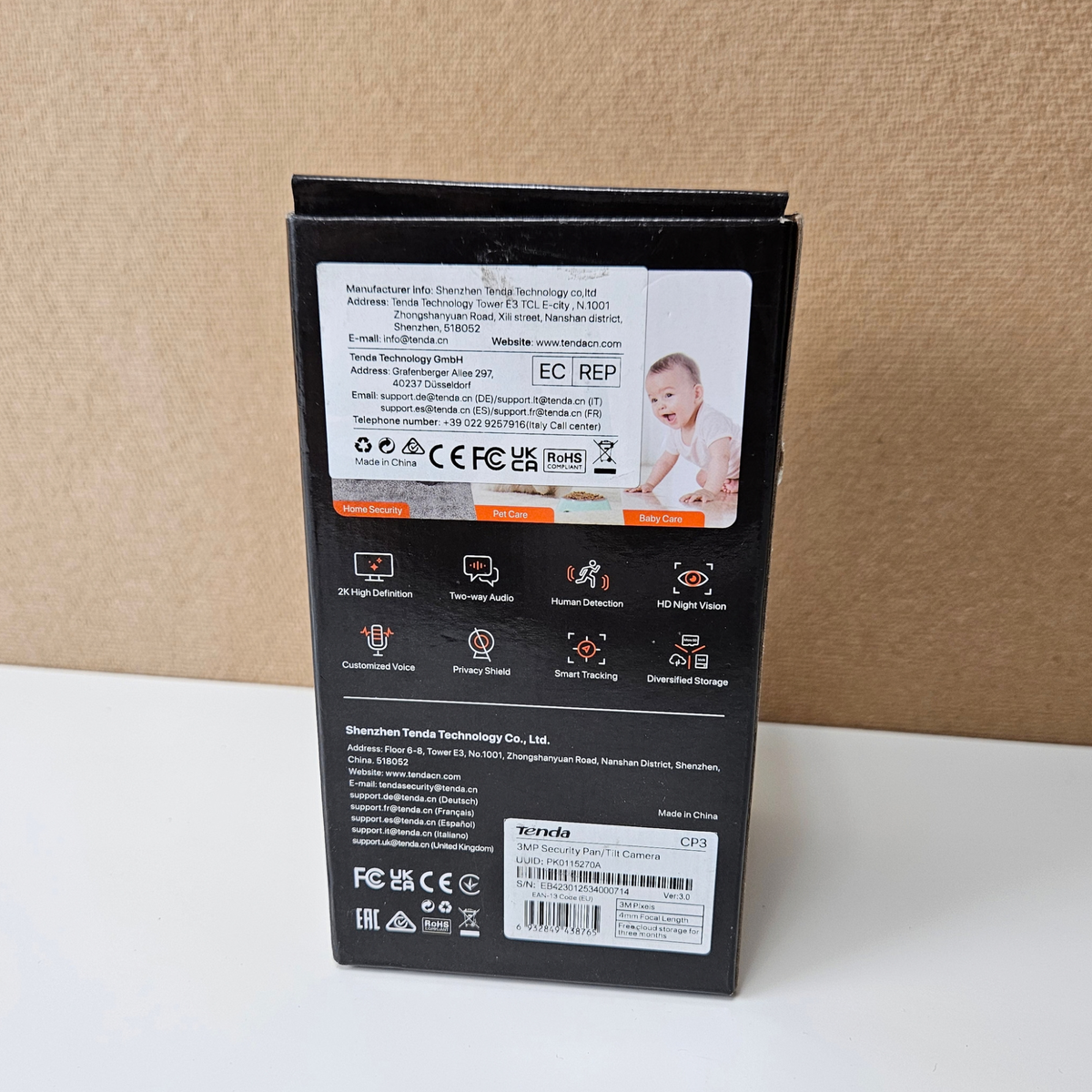 The back of a black retail box, showing technical specifications, icons, and a small photo of a baby, standing on a white surface against a beige textured background.