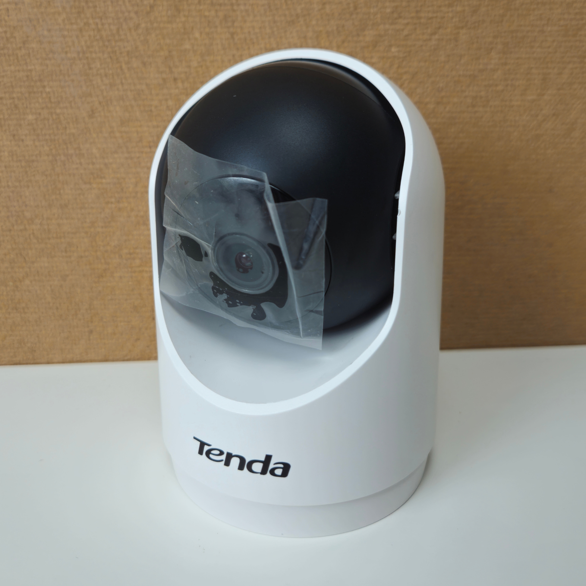 A Tenda dome security camera with a white base and black top, featuring a protective clear plastic sleeve over the lens area, set against a brown textured background.
