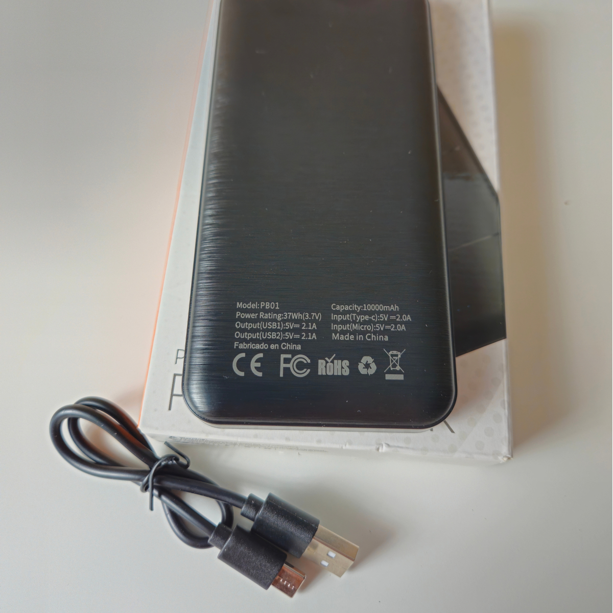 A black rectangular portable power bank with printed technical specifications and certification logos on its back, shown on top of its white retail box with a black USB cable in front on a light surface.