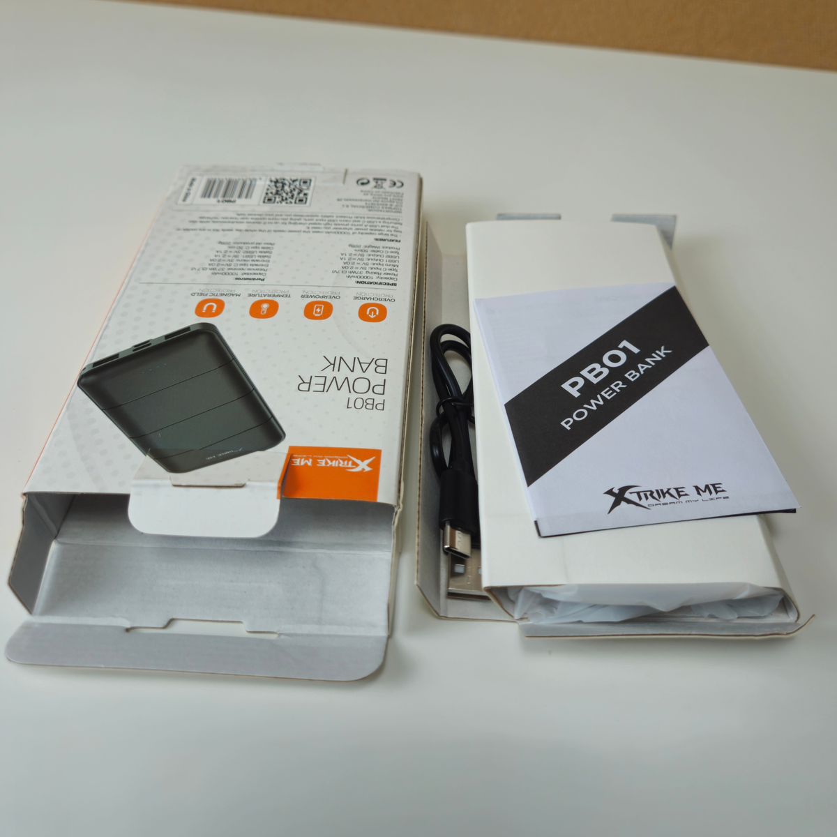 Xtrude Me PB01 Power Bank retail packaging shown open on a white surface, alongside a black USB cable and a small printed booklet. The box includes a photo of the power bank and branding text.