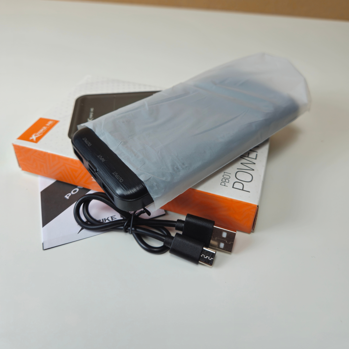 A black portable power bank wrapped in a translucent protective sleeve, shown with its orange and white retail box and a black USB-C to USB-A cable on a light surface.
