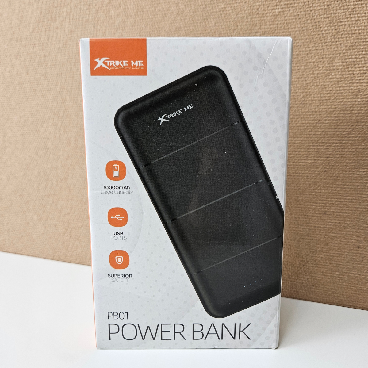 Xtrike Me PB01 Power Bank retail packaging box, showing a black power bank graphic on a white box with orange accents, set against a textured beige background.