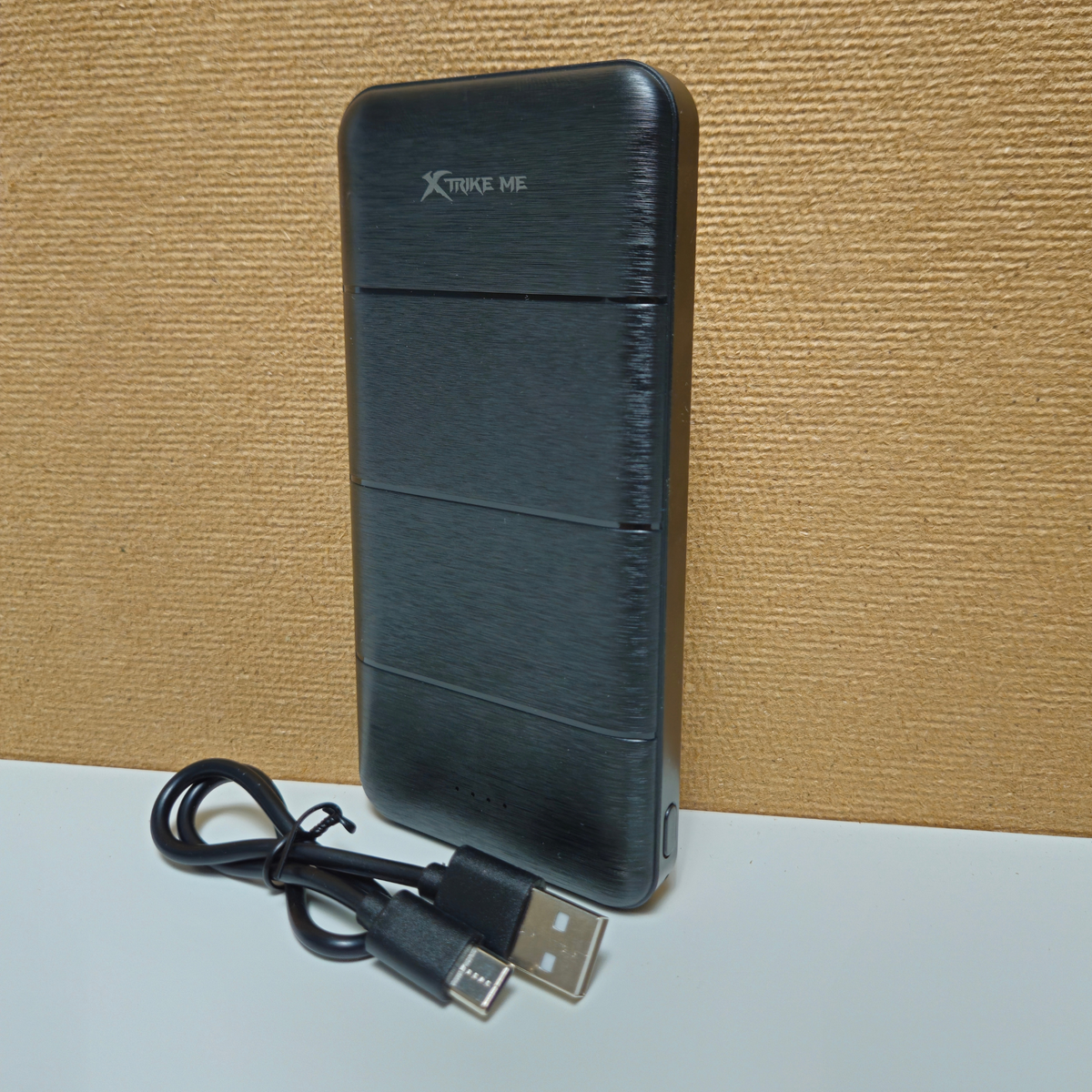 A black brushed-metal portable power bank with a USB-C cable, standing on a white surface against a textured tan background. The power bank has 'Xtrike me' printed on the top.