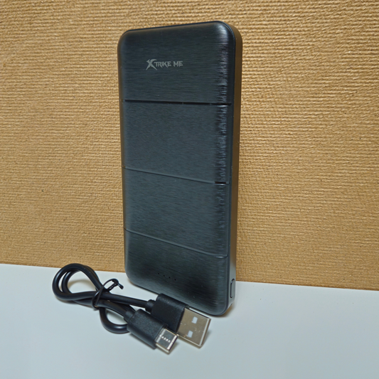 A black brushed-metal portable power bank with a USB-C cable, standing on a white surface against a textured tan background. The power bank has 'Xtrike me' printed on the top.