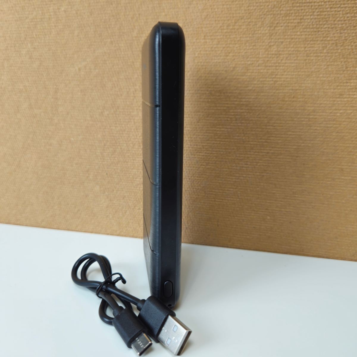 A slim black rectangular electronic device standing upright on a white surface, with a black USB cable placed in front of it against a tan textured backdrop.